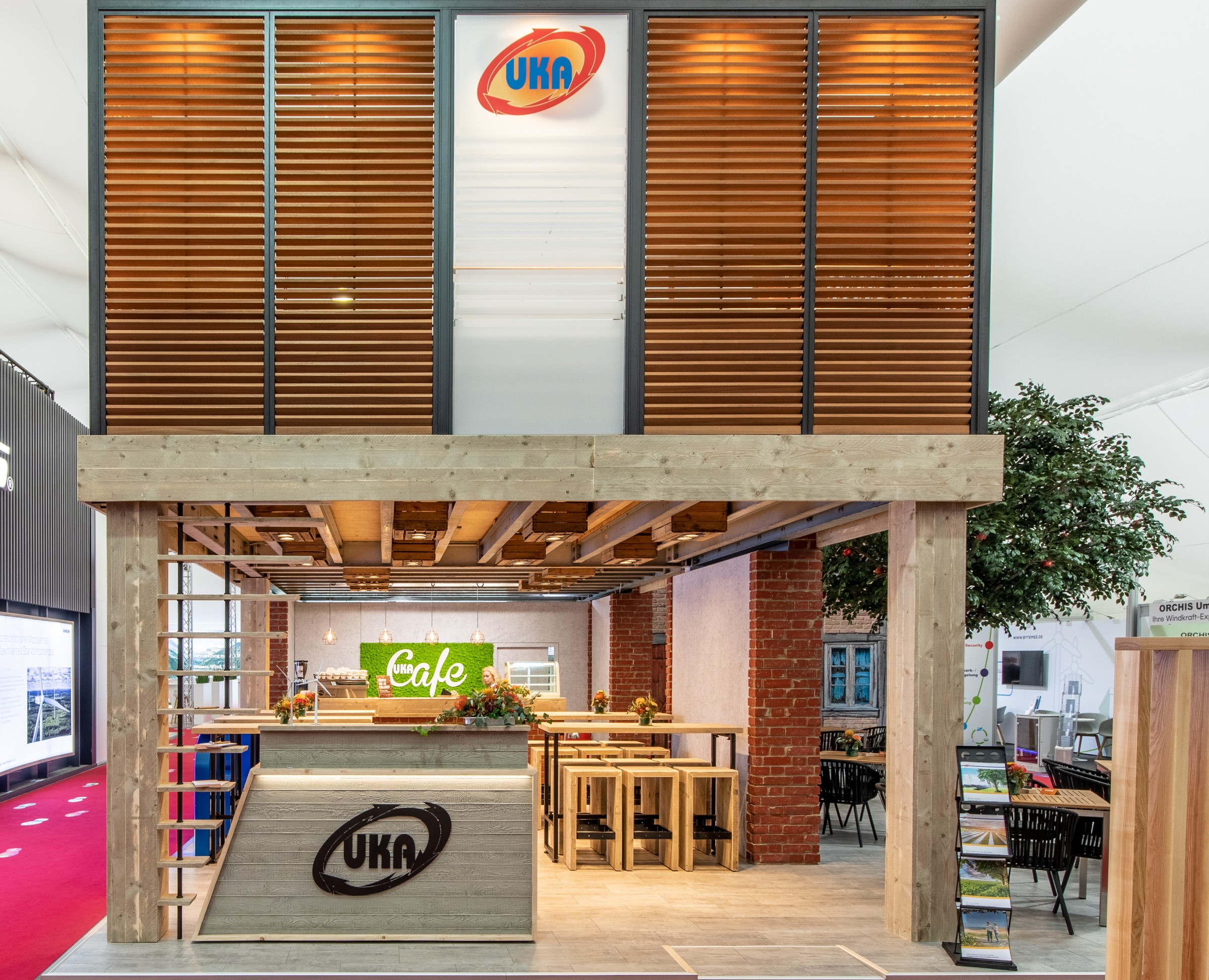 Messestand von UKA mit zweistöckiger Holzkonstruktion, Café-Bereich mit Apfelbaum und grüner ‚Café‘-Leuchtschrift.