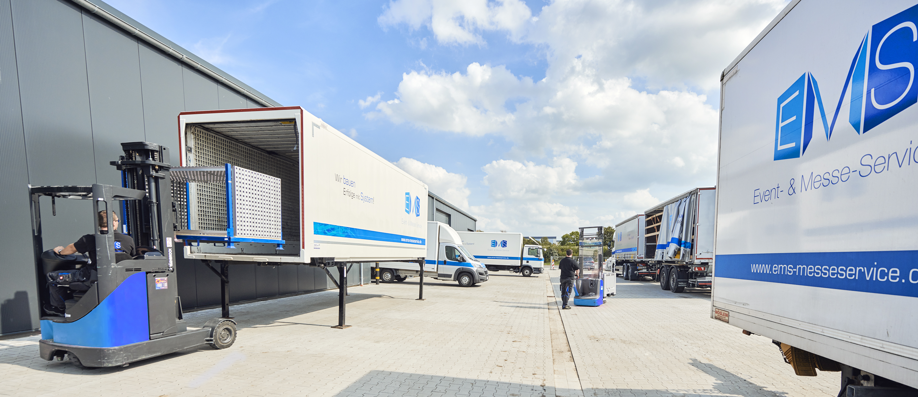Gabelstapler belädt einen EMS-Anhänger mit modularen Messestand-Elementen vor einer Lagerhalle