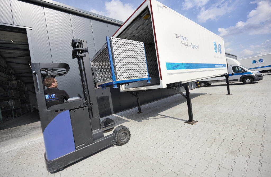 Gabelstapler lädt ein modulares Messestand-Element in einen EMS-Anhänger vor einem Lagergebäude